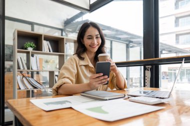 Ofiste dizüstü bilgisayarla çalışırken cep telefonu kullanan mutlu bir iş kadını. Gülümseyen iş kadını akıllı telefonuyla mesajlaşıyor. Güzel stil sahibi bir iş kadını cep telefonunda geziniyor..