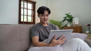 Dijital tablet kullanarak kanepede oturan sıradan giysili çekici Asyalı adam. Teknoloji, iletişim ve yaşam tarzı kavramı.