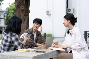 Üniversite kampüsünde otururken bilgisayardan ve tabletten sonuçları kontrol eden bir grup mutlu öğrenci, eğitim, teknoloji ve proje çalışmaları tartışması.