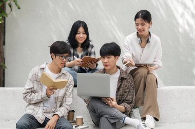 Üniversite kampüsünde otururken bilgisayardan ve tabletten sonuçları kontrol eden bir grup mutlu öğrenci, eğitim, teknoloji ve proje çalışmaları tartışması.