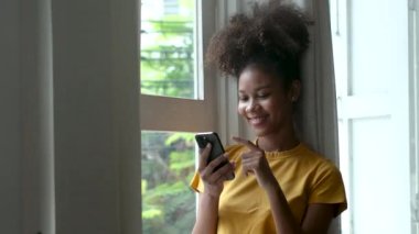 şık siyah kadın akıllı telefon kullanıyor evde mesajlar alıyor rahat yaşam tarzının tadını çıkarıyor pencereden dışarı bakıyor Afro-Amerikan kadın cep telefonuyla mesajlaşıyor.