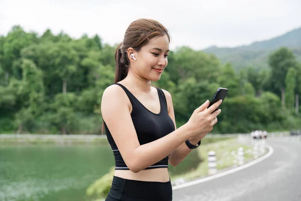 快乐的亚洲女孩运行和检查她的数据 每天的目标在智能手机应用 年轻女子慢跑和看移动屏幕连接耳机播放音乐 — 图库照片