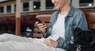 Gezgin genç Asyalı adam tren istasyonunda cep telefonu rezervasyonu yapıyor. Akıllı telefon arama konumu kullanarak trenle iyi yolculuklar. Platform demiryoluna erkek sırt çantalı geldi..