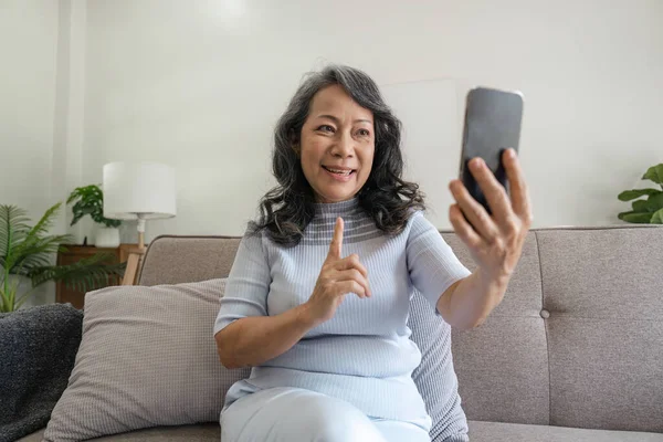 Happy mature middle aged woman waving hand holding mobile phone video conference calling by social distance virtual family online chat meeting sitting on couch at home.
