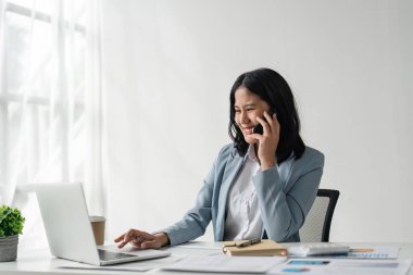 Çekici genç iş kadını cep telefonuyla konuşuyor ve iş yerinde ve dizüstü bilgisayarında otururken gülümsüyor..