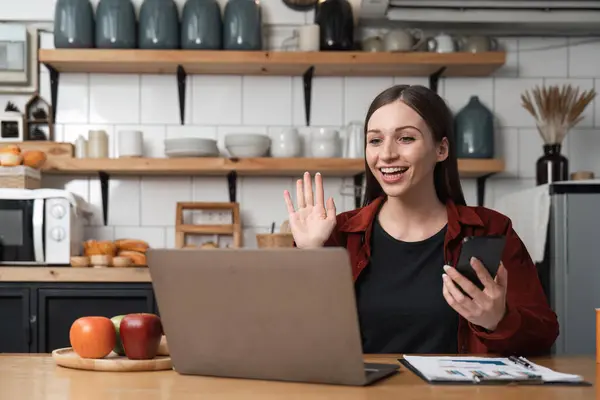 Uśmiechnięta Młoda Kobieta Rozmowie Wideo Kolegami Siedząca Kuchni Laptopem Machająca — Zdjęcie stockowe