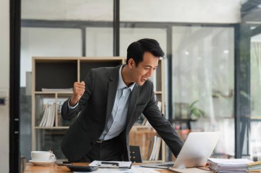 Heyecanlı iş adamı başarıyı kutluyor, e-postada iyi haberler okuyor, mutlu aşırı mutlu iş adamı dizüstü bilgisayara bakıyor, evet işareti yapıyor ve gülüyor, çalışma masasında oturuyor..