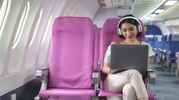 キャビンインテリアでの旅行中に音楽を聞いて ネット上でオンラインウェブサイトをチャットイヤフォンで肯定的な女性旅行ブロガー 飛行中の技術を使用している若い女性の飛行機の乗客 — ストック動画