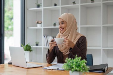 Asyalı Müslüman iş kadını elinde kahve fincanıyla ofiste oturmuş pencereden dışarı bakıyor. iş adamları, çeşitlilik ve ofis kavramı.