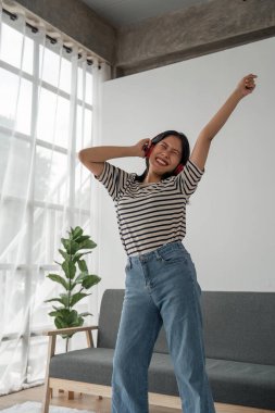 Müzik uygulaması dansıyla müzik dinleyen çekici, mutlu, Asyalı genç bir kadın. Gri kanepenin yanında el kaldırın. Evde dinlenin..