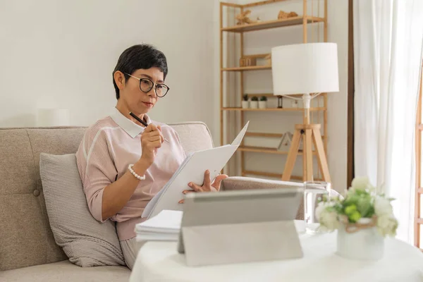 stock image Confident elderly businesswoman is doing paperwork sitting with a tablet on the couch at home.