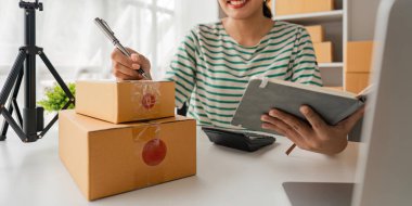 Asyalı bir kadın bir e-ticaret şirketi, onlara göndermeden önce müşterilerin bir listesini kağıda yazıyor. Web sitelerinde ve sosyal medyada bir e-ticaret şirketi işletiyor. Çevrimiçi ürün satma kavramı.