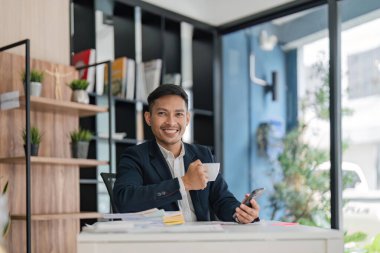 Bilgisayarlı, cep telefonlu, kahveli Asyalı yakışıklı adam ofiste mutlu ve gülümsüyor..