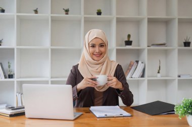 Asyalı Müslüman iş kadını elinde kahve fincanıyla ofiste oturmuş pencereden dışarı bakıyor. iş adamları, çeşitlilik ve ofis kavramı.