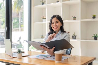 Ofiste dizüstü bilgisayarda çalışan genç ve mutlu bir iş kadını masa başında kameraya bakıyor, iş yerinde kadın portresi yapıyor..