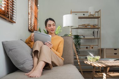 Gri kanepede oturan ve sabahları oturma odasında dijital tablet kullanan güzel ve çekici Asyalı bir kadın..