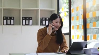 Gülümseyen genç iş kadını telefonda tablet kullanarak konuşuyor. Ofis masasına otur..