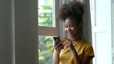 şık siyah kadın akıllı telefon kullanıyor evde mesajlar alıyor rahat yaşam tarzının tadını çıkarıyor pencereden dışarı bakıyor Afro-Amerikan kadın cep telefonuyla mesajlaşıyor.