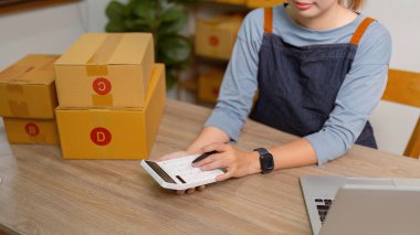 KOBİ sahibi, Asyalı kadın girişimci, çevrimiçi siparişleri kontrol ediyor ve ürün fiyatlarını hesaplamak ve ürünleri kutulara koymak için bir fiyat hesap makinesi kullanıyor.