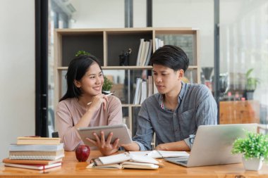 Eğitim, öğretim, öğrenme, teknoloji ve insan kavramı. İki Asyalı erkek ve bayan sınıf arkadaşı sınıf arkadaşlarının ödevlerini yapmalarına yardım ediyor, arkadaşlarıyla ders kitapları okuyorlar..
