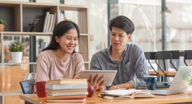 Eğitim, öğretim, öğrenme, teknoloji ve insan kavramı. İki Asyalı erkek ve bayan sınıf arkadaşı sınıf arkadaşlarının ödevlerini yapmalarına yardım ediyor, arkadaşlarıyla ders kitapları okuyorlar..