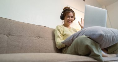 Asyalı kadın, oturma odasındaki kanepede otururken dizüstü bilgisayarla uzaktan iletişim kurarak el sallıyor..