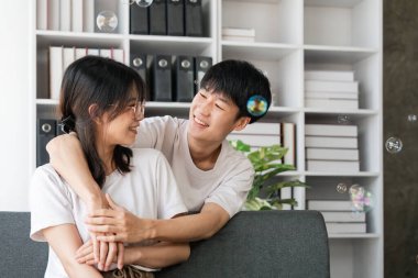 Mutlu genç Asyalı çift rahat rahat evdeki kanepede oturup sarılıyorlar birbirlerine bakıyorlar, hafta sonunun keyfini çıkarıyorlar..