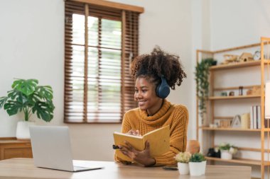 Uzaklık Eğitimi. Gülümseyen Afro-Amerikan kadın portresi masada oturuyor, dizüstü bilgisayar kullanıyor ve deftere yazıyor, not alıyor, özel ders, konferans ya da webinar izliyor, evde internetten ders çalışıyor..