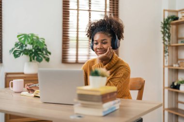 Uzaklık Eğitimi. Gülümseyen Afro-Amerikan kadın portresi masada oturuyor, dizüstü bilgisayar kullanıyor ve deftere yazıyor, not alıyor, özel ders, konferans ya da webinar izliyor, evde internetten ders çalışıyor..