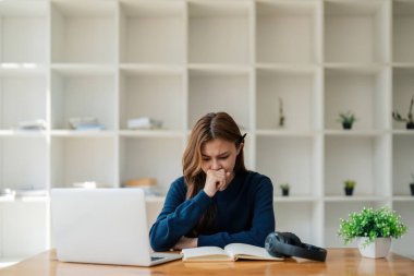 Laptoptan sıkılmış Asyalı bir kadın uzakta öğrenme veya online eğitim konusunda üzgün ve hayal kırıklığına uğramış bir şekilde evde oturuyor..