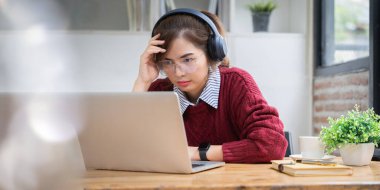 Evde kırmızı tişört giyen Asyalı bir üniversite öğrencisi online üniversite derslerine girip kulaklıkla dinliyor..