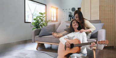 LGBT 'nin iki kız arkadaşı evdeki pencerenin yakınındaki kanepede gitar çalıyorlar. lezbiyen yaşam tarzı aşk kazandı..