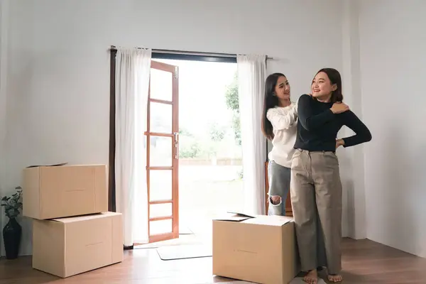 Homosexual happy young Asian woman help massage body pain girlfriend while carrying their belongings relocating into new rent house.