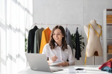 Terzi kadının portresi terzi atölyesinde giysiler üzerinde çalışıyor. Çekici genç bayan moda tasarımcısı atölyede dizüstü bilgisayarda çalışıyor..