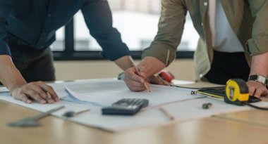 İnşaat planı üzerinde çalışan mühendis ekibinin yakın çekim eli inşaat alanında kaskla birlikte çalışma masasının planını tartışıyorlar..