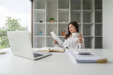 Genç mutlu Asyalı iş kadını ofis bilgisayarında çalışıyor gülümsüyor, kahve içiyor rahatlıyor..