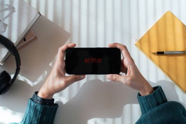 Kulaklıklı Netflix uygulamalı iPhone 14 Pro akıllı telefonu olan bir kadın. Ofis ışıklarının gölgesi Chiang Mai Tayland. SEPT.05 2023..