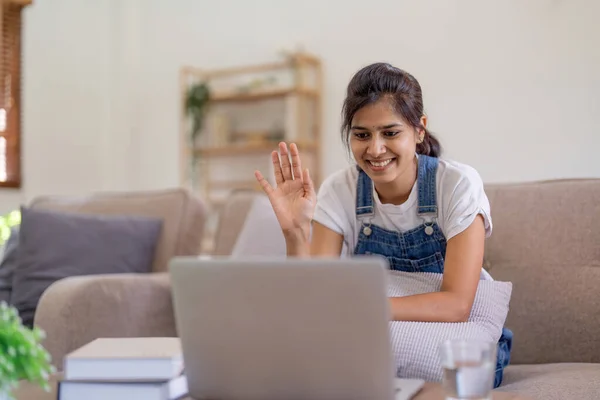 Cheerful young indian woman using laptop for video call with friend or family sitting down on the comfortable couch at home.