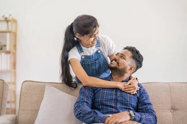 Happy lovely young Indian couple together at home, young wife hugging from behind her husband, sitting and rest on sofa in home, portrait of romantic multiracial couple in love.