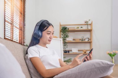 Genç Asyalı kadın evdeki oturma odasında kanepede müzik dinliyor. Mutlu Asyalı kadın cep telefonu kullanıyor, kulaklık takıyor ve kanepede oturuyor..