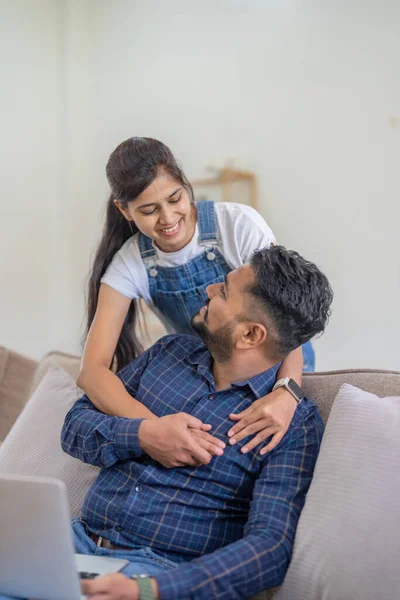 Young indian couple using laptop while sitting on couch and floor. Happy smiling indian woman embrace from behind her boyfriend while watch video on laptop at home.