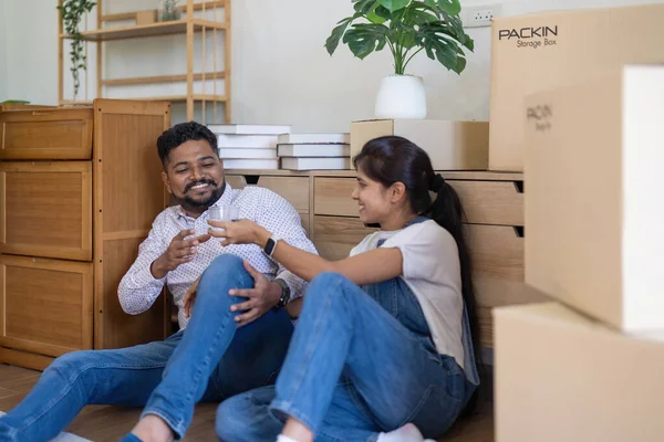 Young indian couple taking a break on moving day into new home sitting on floor and drinking water surrounded by removal box.