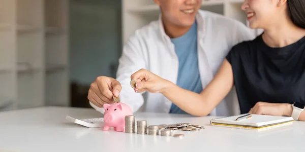 Family savings. Happy couple asian putting coin in piggybank while sitting together on desk at home, loving spouse planning budget and saving money for future.