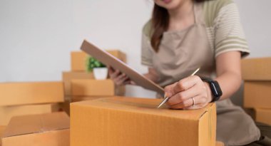 Asyalı kadın, onlara göndermeden önce kağıda bir müşteri listesi yazıyor. Web sitelerinde ve sosyal medyada ekomünikasyon işi yapıyor. Çevrimiçi ürün satma kavramı.