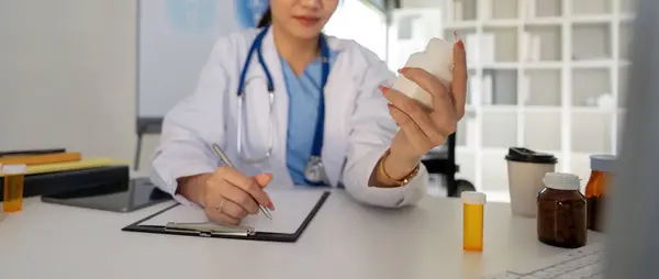 Female doctor writing in a notebook and holding a medicine bottle works at a computer while the table while sitting at a work desk.