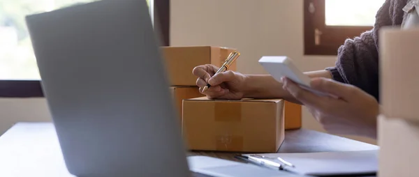 Woman is writing a list of customer on box before shipping to them, she runs an ecommerce business on websites and social media. Concept of selling products online.