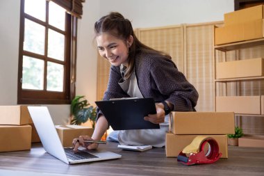 Girişim KOBİ küçük işletme girişimcisi. İnternetten sipariş almak ve gözden geçirmek için dizüstü bilgisayar ve kutu kullanan serbest Asyalı kadın müşterilere, çevrimiçi iş fikirlerine satış hazırlamak için internetten sipariş alıyor ve gözden geçiriyor..