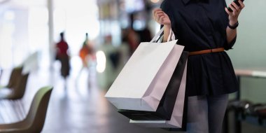 Akıllı telefon kullanan bir kadın alışveriş merkezinin arka planında merdivenlerde dururken Kara Cuma alışveriş çantası tutuyor..