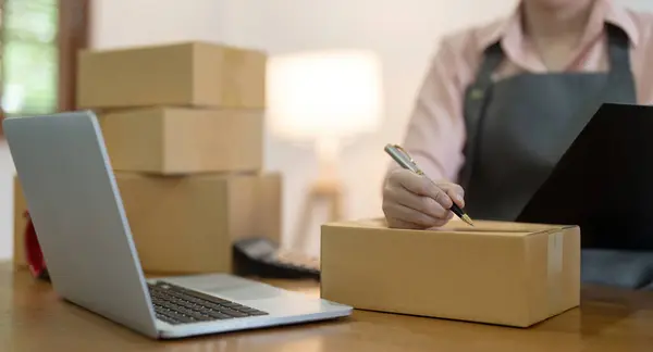 Woman is writing a list of customer on box before shipping to them, she runs an ecommerce business on websites and social media. Concept of sell product online.