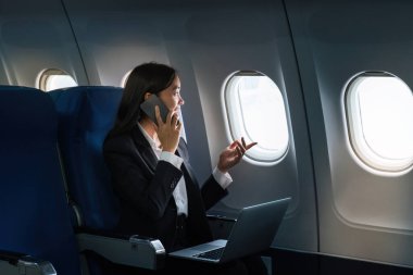 Başarılı Asyalı iş kadını, uçakta dizüstü bilgisayarla çalışan iş kadını ve telefonla konuşurken pencereden dışarı bakan..